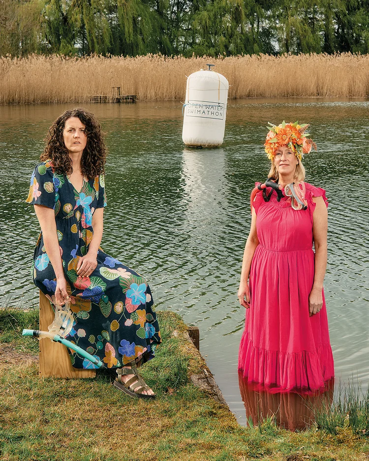 Zwei Frauen stehen am Ufer eines Sees. Die linke Frau trägt ein dunkelblaues Kleid mit buntem Blumenmuster. Die rechte Frau trägt ein rosa Kleid.