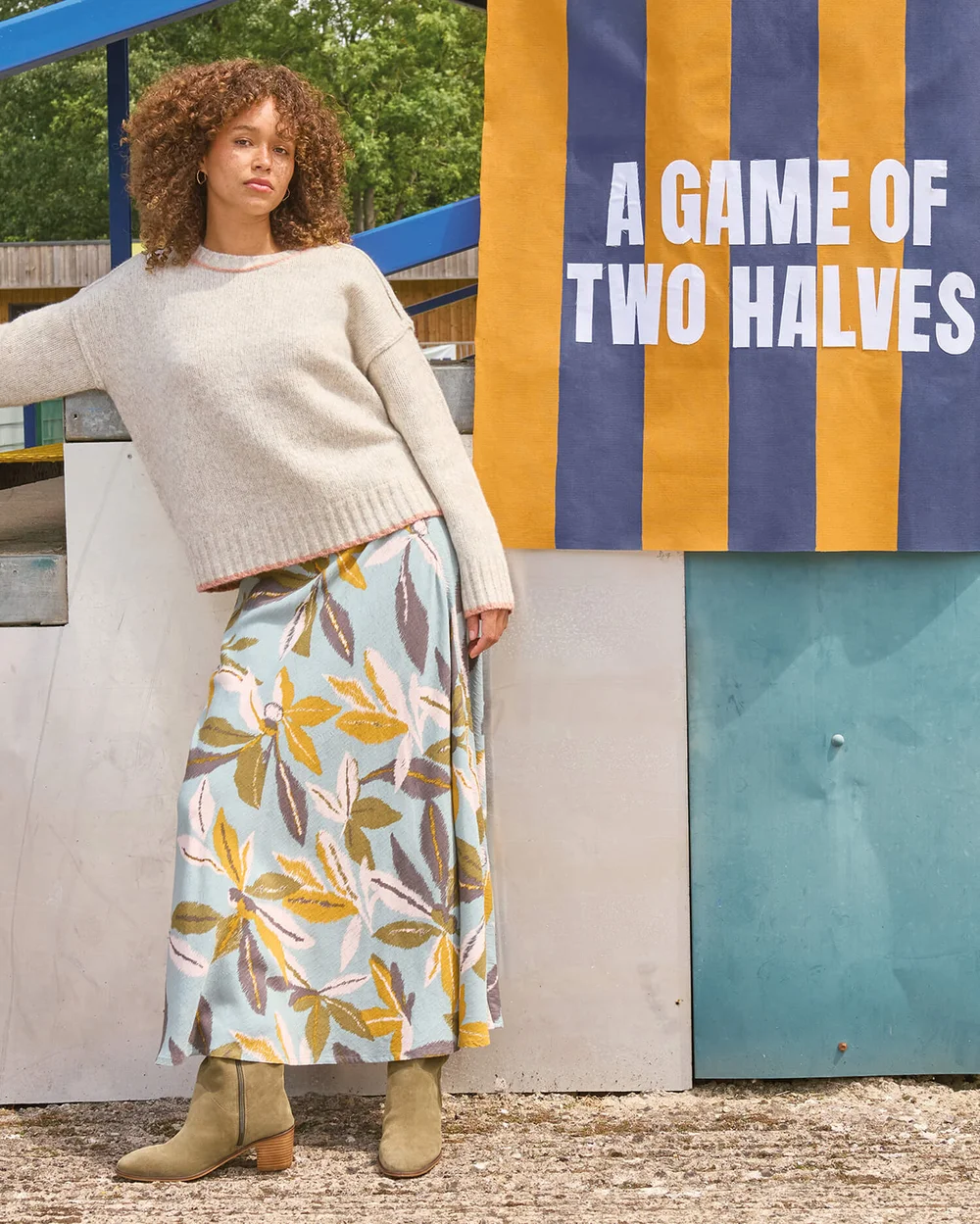 Links im Bild ist eine Frau in einem farbenfrohen Rock und einem Pullover zu sehen, mit einem Banner, auf dem ‚A Game of Two Halves‘ steht.