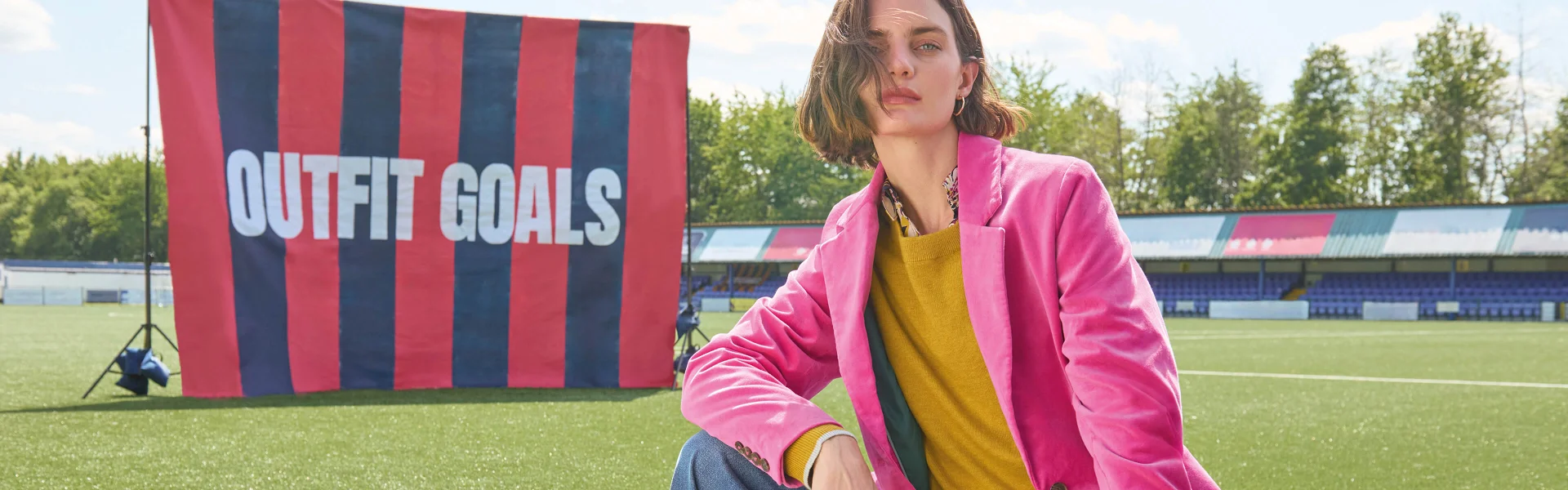 Frau im pinkfarbenen Blazer und gelben Pullover, hinter ihr eine gestreifte Fahne mit der Aufschrift ‚Outfit Goals‘
